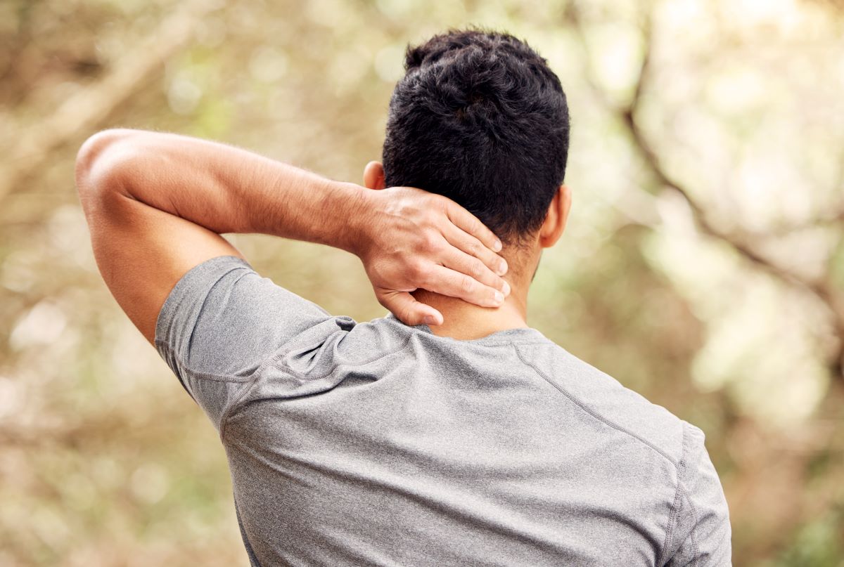 Head-Neck-Masses-symptoms Man in an outdoor setting holding the back of his neck with one hand. The image is from behind and shows the mans back and back of head.