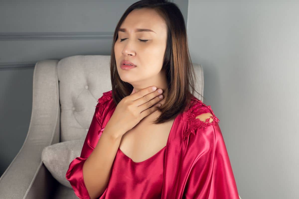 Symptoms of laryngitis and hoarseness Woman at home on corner chair has her hand resting on her upper chest and lower throat area. Her eyes are closed and she appears to be in distress.